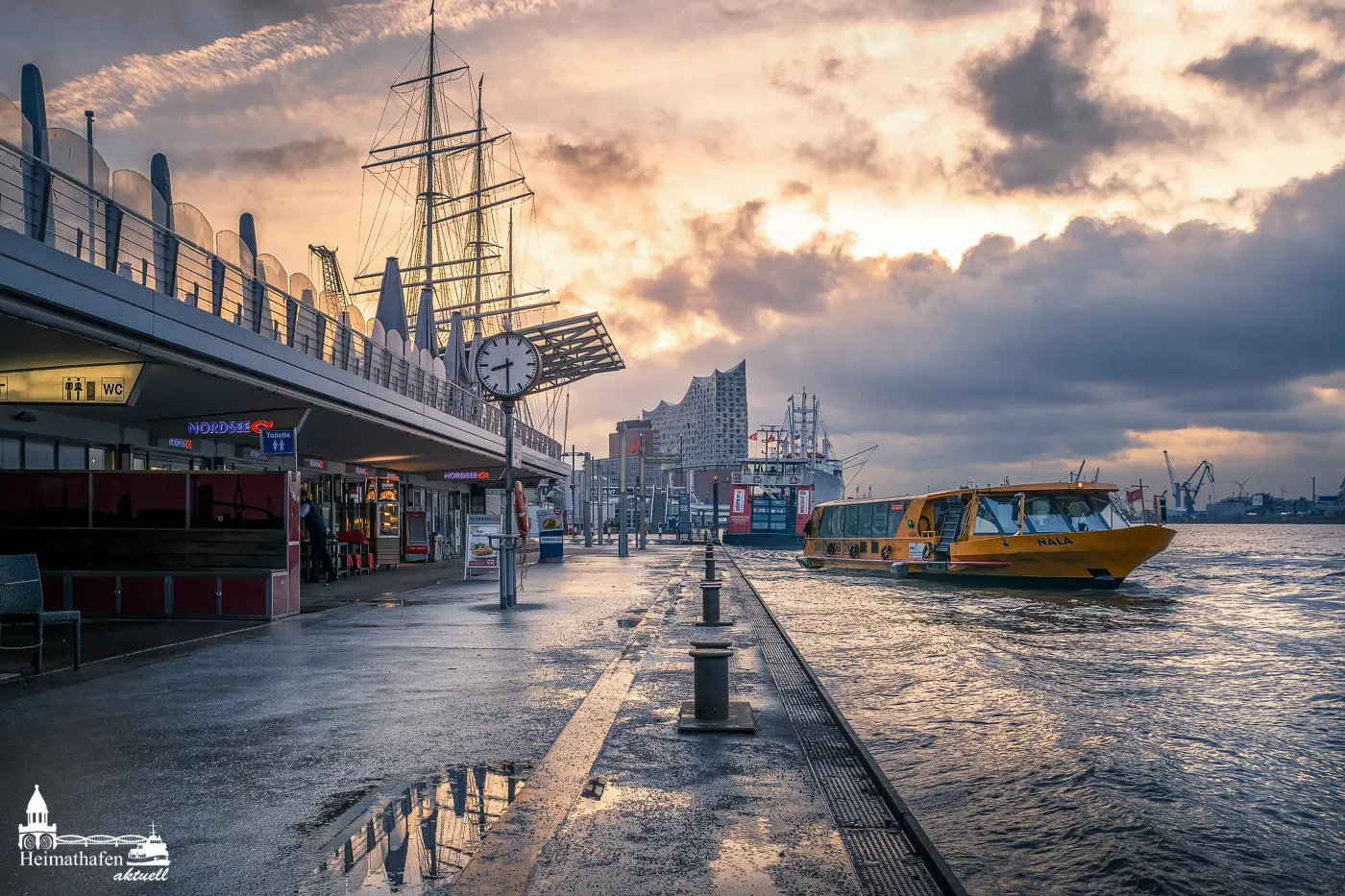 Die Hamburger Landungsbrücken bei dramatischem Himmel, mit Blick auf die Elbphilharmonie und einem gelben Hafenboot.