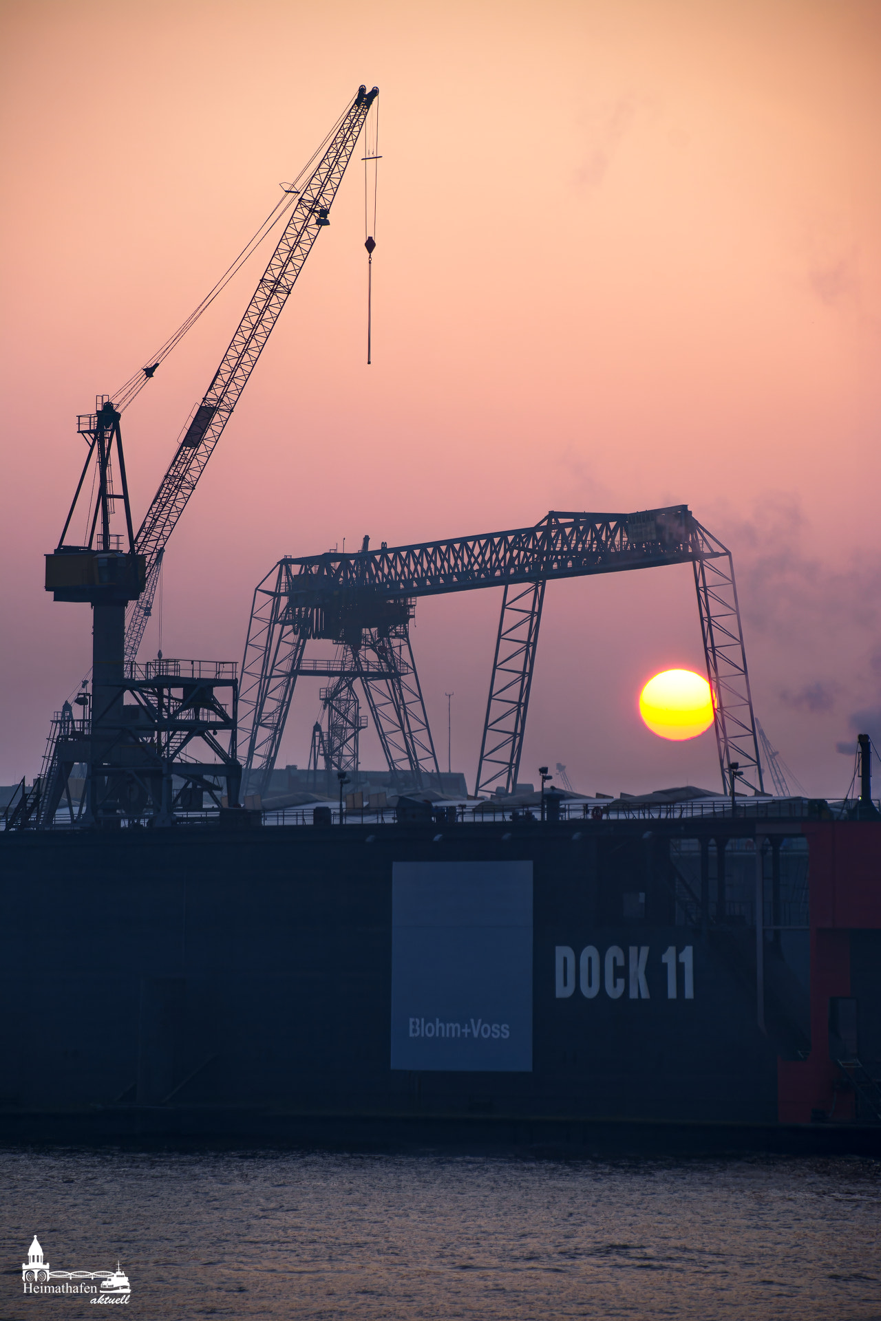 Die aufgehende Wintersonne leuchtet hinter den Hafenkränen von Blohm+Voss am Dock 11 – eine stimmungsvolle Szene voller Industriepracht und Morgentau.