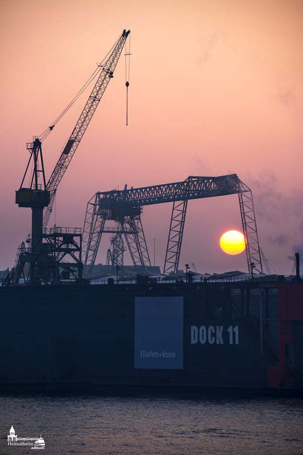 Die aufgehende Wintersonne leuchtet hinter den Hafenkränen von Blohm+Voss am Dock 11 – eine stimmungsvolle Szene voller Industriepracht und Morgentau.
