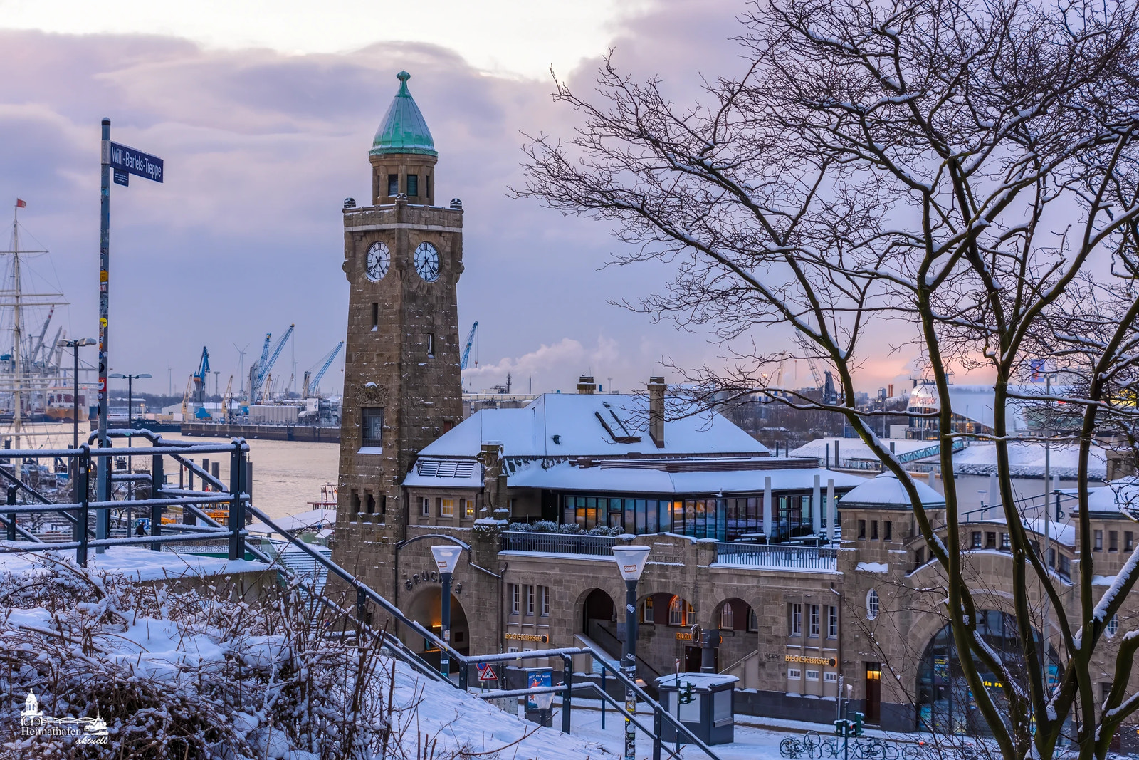 Der Pegelturm der Hamburger Landungsbrücken im Winter, umgeben von verschneiten Dächern und Bäumen. Im Hintergrund ragen Kräne am Hafen in den kalten Himmel.