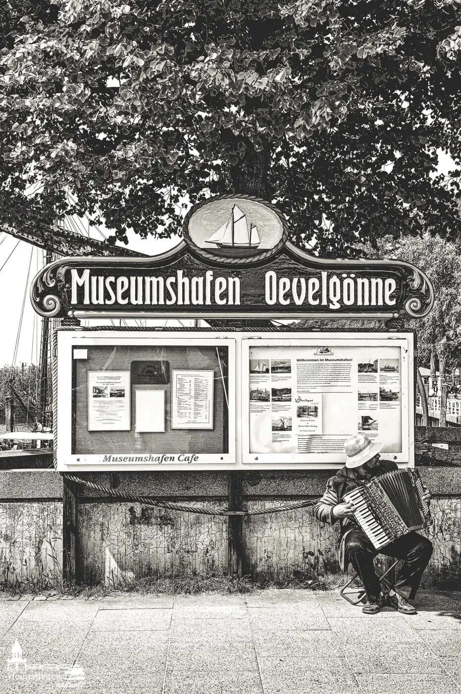 Schwarzweißfoto vom Museumshafen Oevelgönne mit Akkordeonspieler vor Infotafel